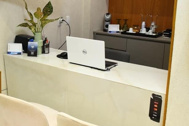 Balcão de recepção em mármore claro, com computador, agenda e uma planta ao lado.